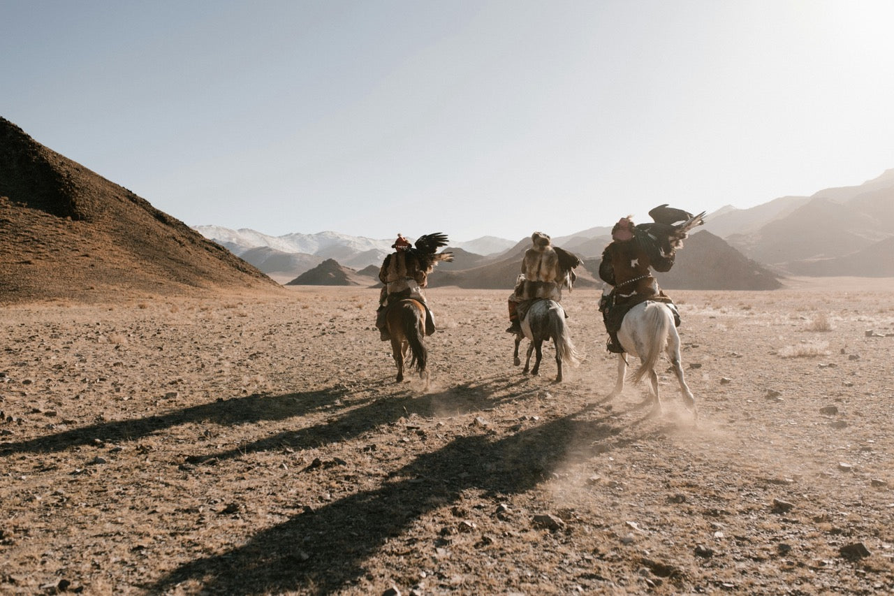 Four people riding horses across a desert landscape with mountains in the background.